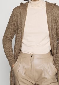 Hnědý pletený kardigan s kapucí, doplněný světlobéžovým rolákem a koženými plisovanými kalhotami v barvě bělené kůže, vytvářející texturovaný vrstvený vzhled.