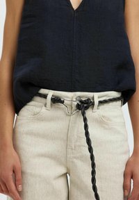 Ceinture tressée noire avec quincaillerie en acier, portée sur un pantalon texturé beige clair taille haute, associée à un haut sans manches sombre.