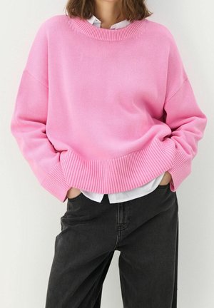 Kobieta ubrana w luźny różowy sweter z dzianiny na białej koszuli, czarne wysokie dżinsy z wysokim stanem, ręce w kieszeniach, stoi na tle białej ściany.