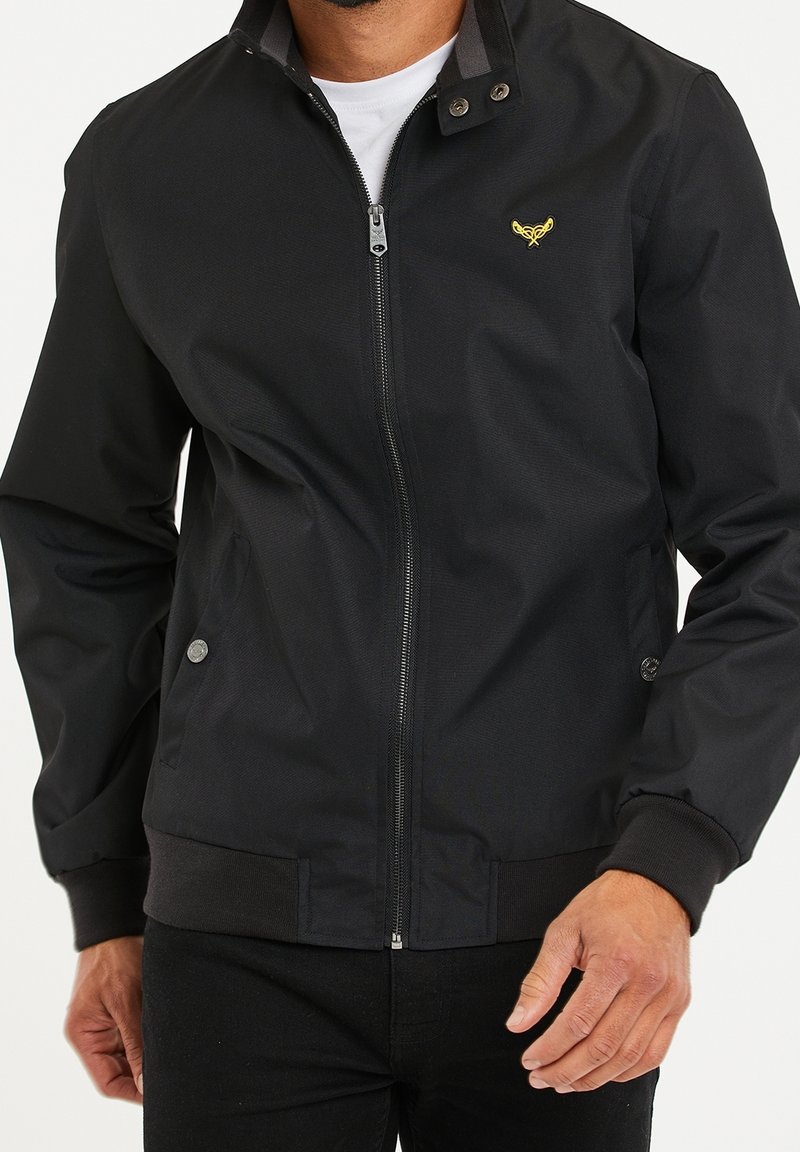 Homme portant une veste noire à fermeture éclair avec poignets et ceinture côtelés, et un petit logo brodé jaune sur la poitrine, par-dessus une chemise blanche.