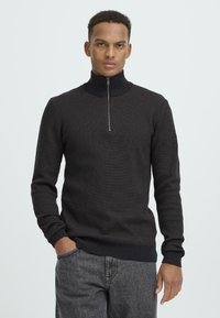 Tmavosivý pletený sveter s vysokým golierom a polovičným zipsom, s dlhými rukávmi a ribovanými manžetami a spodným lemom. Nosí sa s sivými nohavicami.