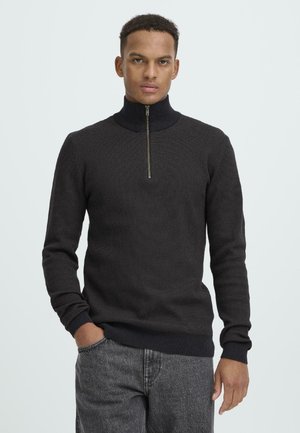 Dunkelgrauer Strickpullover mit hohem Kragen und halbem Reißverschluss, der lange Ärmel sowie gerippte Bündchen und Saum aufweist. Getragen mit grauen Hosen.
