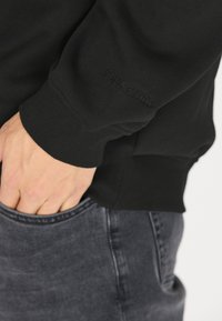 Mustapaita, jossa on joustavat hihansuut ja vyötärö, ja hiha on varustettu pienellä kohokuvioidulla logolla. Paita on yhdistetty tummiin farkkuihin, joissa on etutasku.