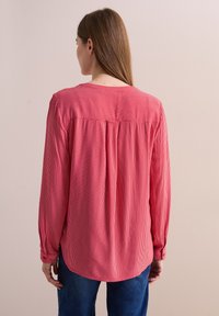 Blusa a maniche lunghe a righe rosse con collo rotondo, tessuto testurizzato e dettaglio plissettato sul retro, indossata con pantaloni in denim.