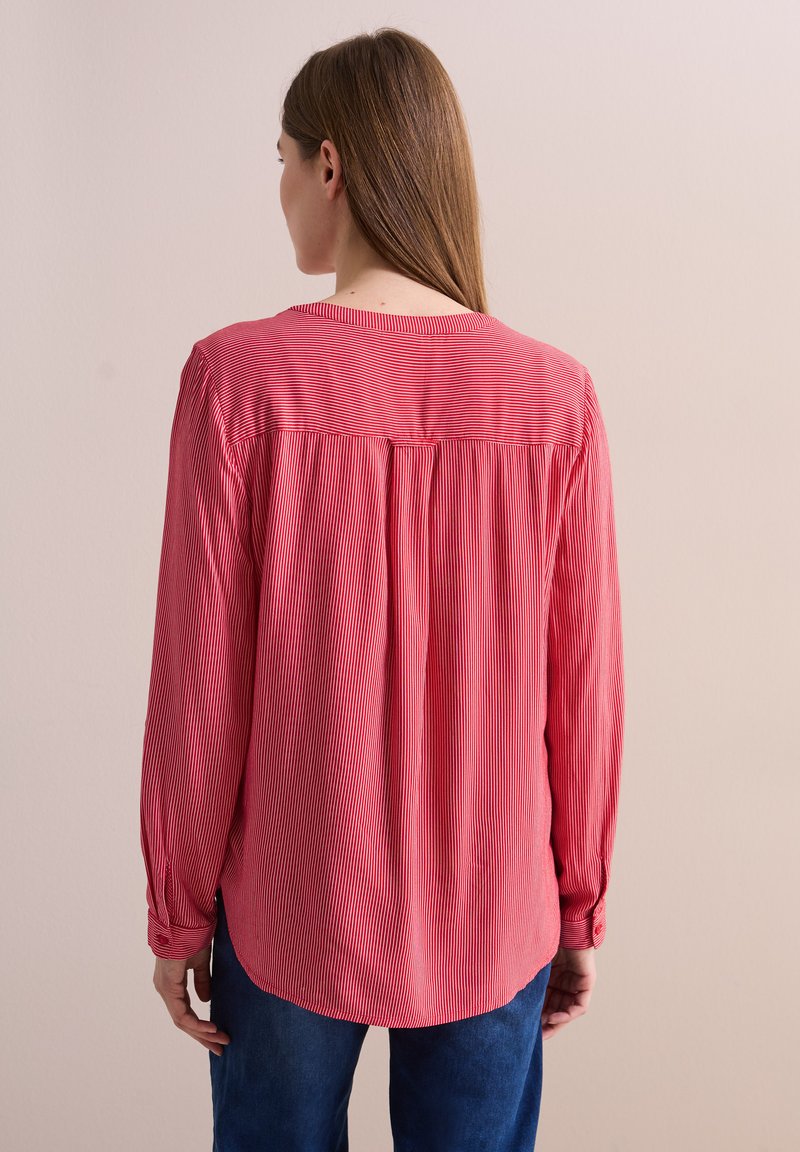 Blusa a maniche lunghe a righe rosse con collo rotondo, tessuto testurizzato e dettaglio plissettato sul retro, indossata con pantaloni in denim.