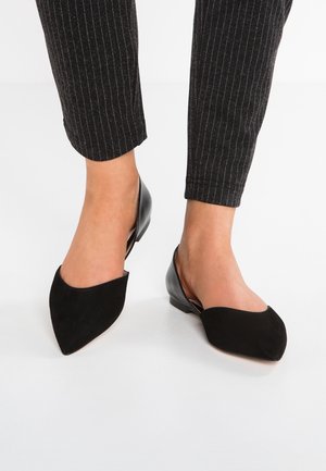 Pieds portant des ballerines slingback noires à bout pointu associées à un pantalon noir à rayures fines, longueur cheville, sur un sol blanc.