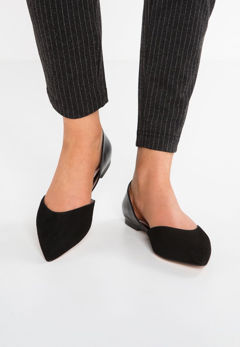 Pieds portant des ballerines slingback noires à bout pointu associées à un pantalon noir à rayures fines, longueur cheville, sur un sol blanc.