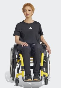 Juoda sportinė marškinė su balta logotipu, juodos kelnės ir juodi batai. Asmuo sėdi geltonoje neįgaliųjų vežimėlyje su metaliniais akcentais.