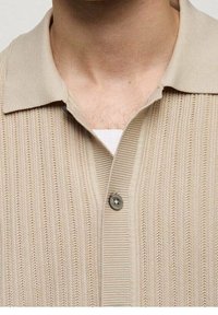 Camisa polo de punto en color beige con un patrón texturizado. Presenta un cuello corto y un cierre de un solo botón en el cuello.