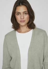 Světlý zelený cardigan s výstřihem do V, vyrobený z měkkého materiálu. Má dlouhé rukávy a volnější střih, který je přístupný přes bílý top.