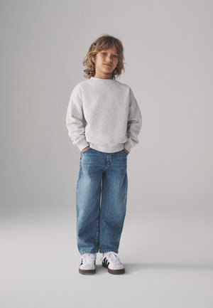 Barn med krøllet hår iført en lysegrå sweatshirt, brede blå jeans og hvide sneakers med sorte striber, står med hænderne i lommerne.