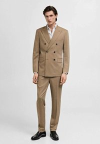 Completo beige a doppio petto con rever a scasso, tasche frontali e bottoni neri. Abbinato a una camicia a righe chiare e scarpe nere lucide.