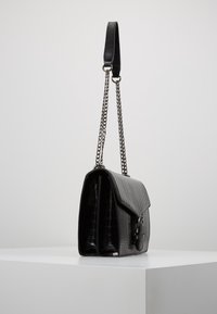 Schwarze Lederhandtasche mit einer strukturierten Krokodilprägung, ausgestattet mit einem Kettenriemen und einem Klappenverschluss, aufrecht auf einer weißen Fläche präsentiert.