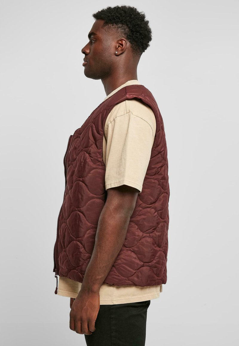 Gilet matelassé bordeaux, sans manches, avec une fermeture éclair à l'avant, motif texturé, porté sur un t-shirt beige.