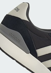 Gros plan sur une basket Adidas noire et blanche montrant un talon texturé, un logo gris sur la semelle et des bandes cousues sur le côté.