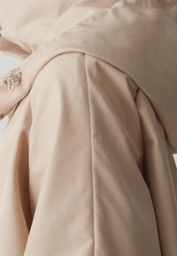 Cappotto impermeabile beige con texture liscia, cappuccio strutturato e chiusura anteriore con toggles, dal design minimalista.