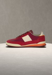 Κόκκινο και μπορντό sneaker χαμηλού προφίλ με μπεζ πλαϊνή πινακίδα, πορτοκαλί λεπτομέρεια στην σόλα και μπορντό κορδόνια, εμφανίζεται στο προφίλ πάνω σε ουδέτερη επιφάνεια.