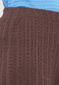 Καφέ υφασμάτινη υφή με ανάγλυφο σχέδιο, διαθέτει εφαρμοστό ζωνάρι μέσης. Το ύφασμα φαίνεται απαλό και ελαφρώς ελαστικό.