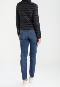 Svart vadderad jacka med horisontell sömnad, blå jeans och vita sneakers. Jackan har hög krage och dragkedja vid ärmslut.