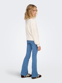 Weißer, flauschiger Pullover mit rundem Ausschnitt, kombiniert mit hellblauen, ausgestellten Jeans und braunen Slippern mit weißen Akzenten.