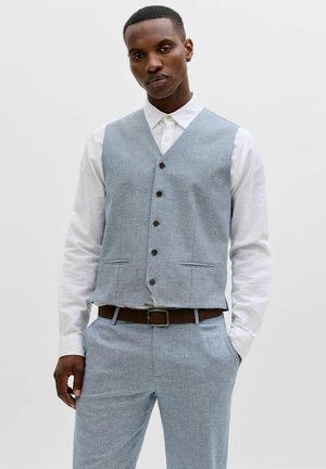 Uomo che indossa un gilet grigio chiaro abbottonato sopra una camicia bianca con pantaloni coordinati e cintura marrone, in piedi con una mano in tasca.