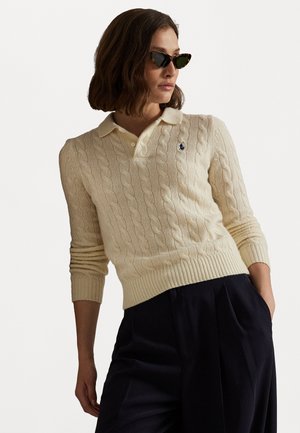 Creme kabelstrikket polo genser med krage, ribbestrikkede mansjetter og hem. Har et lite, brodert logo på venstre bryst.