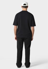Black short-sleeve t-shirt made of cotton, featuring a relaxed fit. Paired with straight black trousers, viewed from the back, with a white cap.