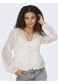 Blusa bianca in cotone con ricami a occhiello, maniche puff lunghe, vita a peplum e un nastro al collo. Indossata sopra una maglietta leggera, abbinata a jeans grigi.