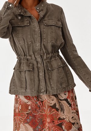 Frau trägt eine olivgrüne, geknöpfte Jacke mit mehreren Taschen und Taillenzug, kombiniert mit einem gemusterten roten und braunen Paisley-Rock.