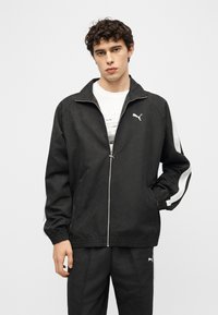 Νέος άνδρας με σγουρά μαλλιά που φοράει μαύρη φόρμα Puma με λευκές ρίγες και λογότυπο, στέκεται με το ένα χέρι στην τσέπη του.