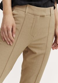 Pantaloni beige su misura con una superficie liscia, dotati di tasche laterali e frontale piatto. La cintura presenta passanti per la cintura e un sottile dettaglio di cucitura.