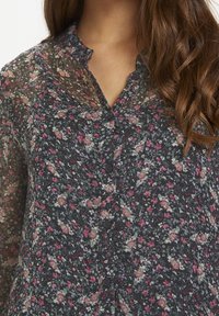 Blumenbluse in dunkler Farbe mit V-Ausschnitt, langen Ärmeln und Knopfverschluss. Der Stoff ist leicht und halbtransparent mit mehrfarbigen Blumenmustern.