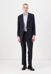 Temno modra dvoredna blazer jakna z zarezanimi klopi, v kombinaciji z ustreznimi krojenimi hlačami in svetlo modro srajco. Črne čevlje.