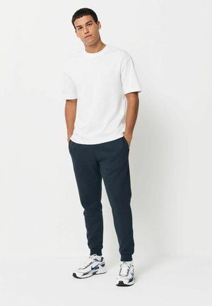 Weißes Kurzarmhemd, dunkelblaue Jogginghose und weiße Sneaker mit schwarzen und blauen Akzenten. Klare Linien und lässige Passform.