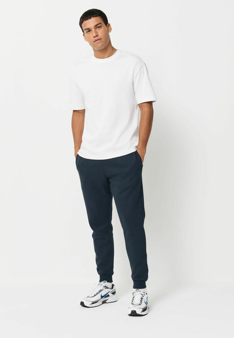 Balta īsā piedurkne krekls, tumši zili sporta bikses un baltas sporta kurpes ar melniem un ziliem akcentiem. Tīras līnijas un brīva piegriezuma.