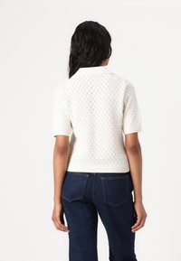 Short-sleeve white knitted sweater with a diamond pattern, featuring a flat collar and ribbed hem. Paired with dark blue jeans.
