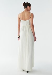 Largo vestido blanco con tirantes finos, con un intrincado borde de encaje y una textura fluida, diseñado con un corsé ajustado y cintura, terminando en un dobladillo festoneado.