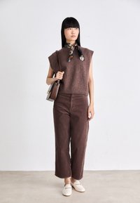 Brown sleeveless knit top, high waist brown wide-leg pants, cream loafers, and patterned scarf tied around the neck, holding a brown handbag.