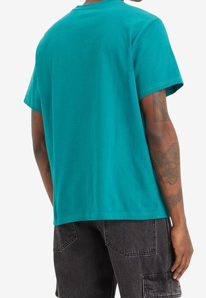 T-shirt en coton turquoise avec un col rond classique et des manches courtes ; présente une texture lisse et un ourlet droit. Pantalon noir visible en dessous.