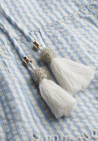 Dos borlas blancas con acentos metálicos y de cuentas unidas a un cordón trenzado de rayas azul y blanca sobre un fondo texturizado.
