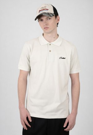 Mladý muž nosící bílé polo triko s vyšitým textem a béžovo-černou truckerskou čepici stojí na světle šedém pozadí.