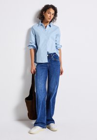 Vrouw in een lichtblauw gestreept blouse, wijdvallende blauwe jeans, witte sneakers, met een donkerbruine leren tas in haar hand, staand tegen een witte muur.