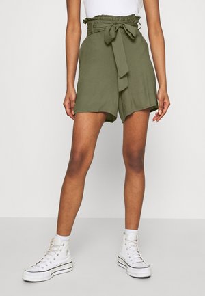Femme portant un short taille haute vert olive avec une ceinture nouée, associé à des chaussettes blanches à la cheville et des baskets montantes blanches.