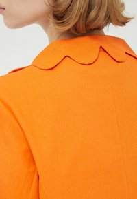 Prenda naranja con cuello escalonado, textura suave y diseño estructurado, destacando un acabado vibrante y de color sólido.