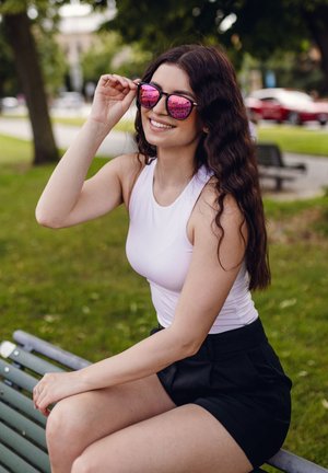 Sluneční brýle s růžovými zrcadlovými skly, černý rám. Model je oblečen v bílém bezrukávovém topu a černých šortkách, sedí na zelené lavičce v parku.