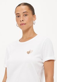 Witte katoenen T-shirt met een klein goudkleurig hartjespatroon op de linkerkant van de borst. Korte mouwen met opgerolde mouwen. Gladde textuur.
