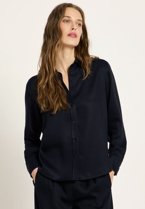 Frau mit langen, welligen Haaren, die ein schwarzes, geknöpftes Hemd und schwarze Hose trägt, steht mit den Händen in den Taschen vor einem schlichten, hellen Hintergrund.