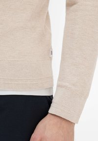 Hellbeiger Strickpullover mit strukturiertem Muster, gerippten Ärmelbündchen und Saum. Enthält seitlich ein kleines Etikett mit der Marke "Blox".