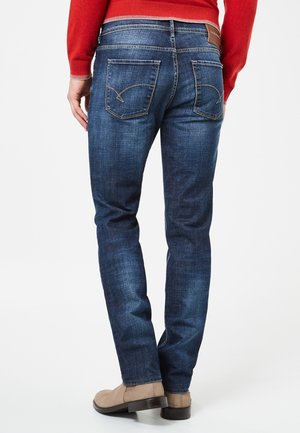 Osoba ubrana w ciemnoniebieskie obcisłe dżinsy, beżowe buty i czerwony sweter, stojąca tyłem do aparatu na białym tle.