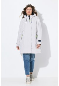 Veste imperméable gris clair avec capuche, présentant des accents noirs et verts, des poches zippées et une coupe décontractée. Portée sur un jean décontracté et des baskets.